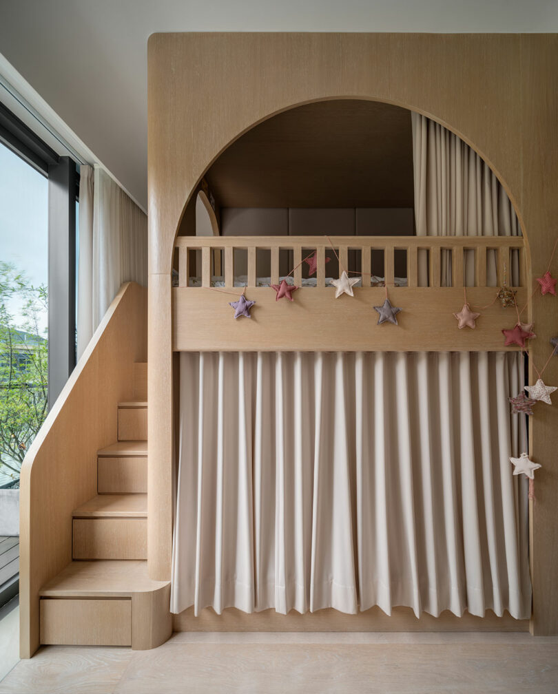 A wooden loft bed with a staircase on the left, white curtains below the bed, and star-shaped decorations hanging in front.