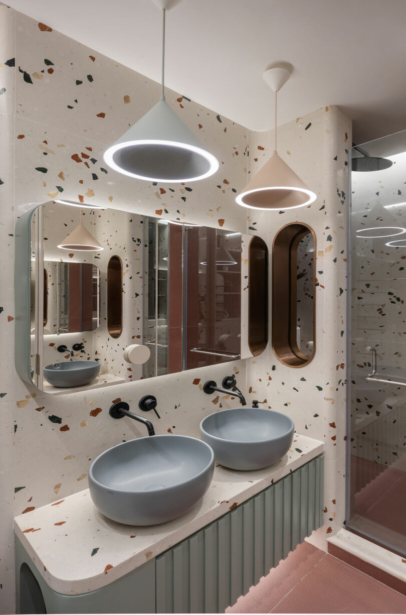 A modern bathroom features terrazzo walls, two round sinks with black faucets, a large mirror, and two pendant lights. A glass shower enclosure is visible in the background.