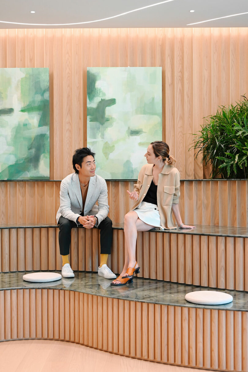 Two people engage in conversation while seated on tiered, wooden seating. The background features abstract, green paintings and a potted plant.