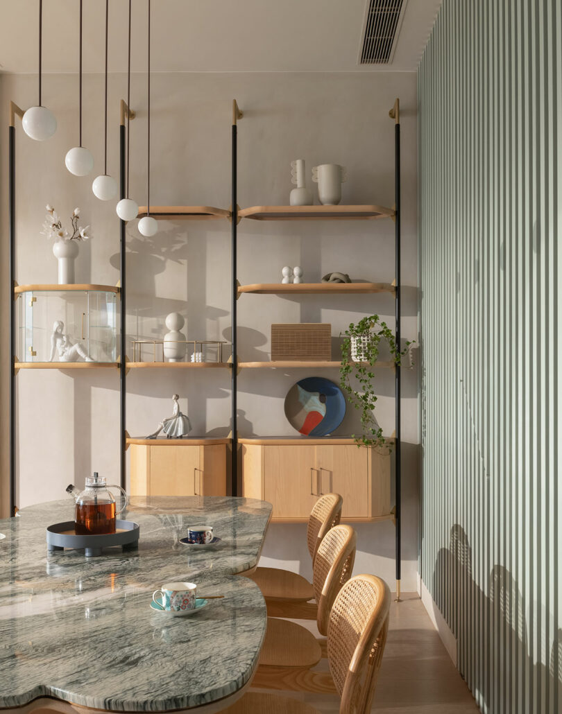 Dining area with a marble table, wooden chairs, a teapot, and a cup. Wooden shelves on the wall hold decorative items and a potted plant. Vertical green paneling is seen on the right.