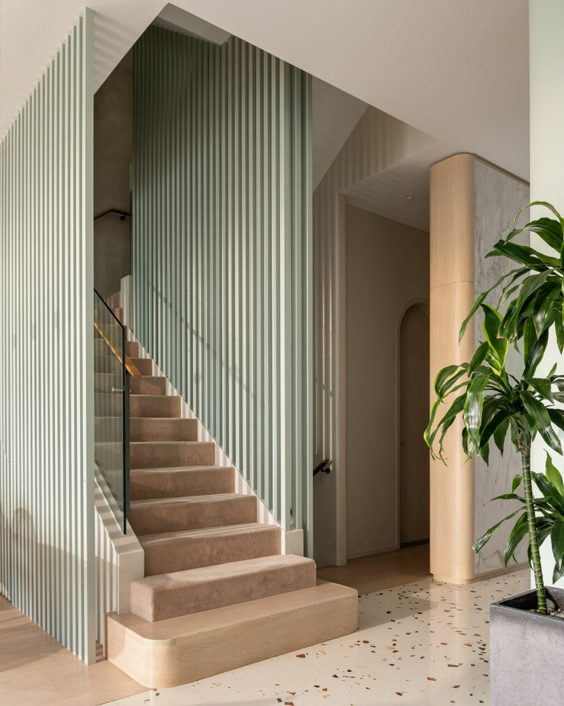 A modern, minimalist staircase with beige carpeting and glass railing, surrounded by vertical slats. Plant near bottom right. Neutral-toned interior with terrazzo flooring.