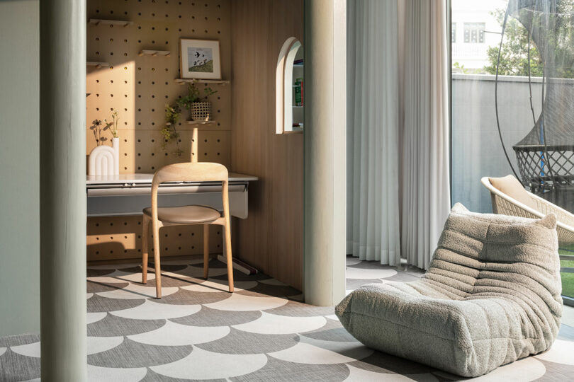 A modern home office with a wooden chair, desk, pegboard wall organizer, arched shelf, and large floor cushion. Floor-to-ceiling windows provide ample natural light.