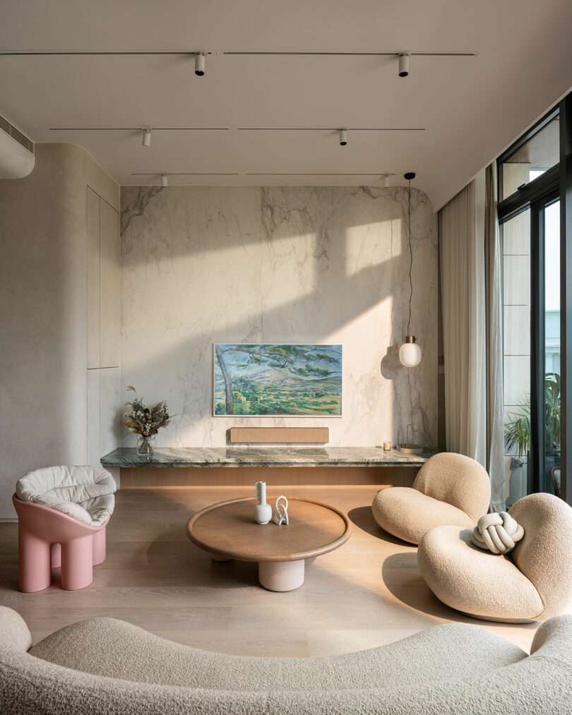 A modern living room with beige furniture, a rounded wood coffee table, and a marble accent wall with a mounted TV. Large windows on the right let in natural light.