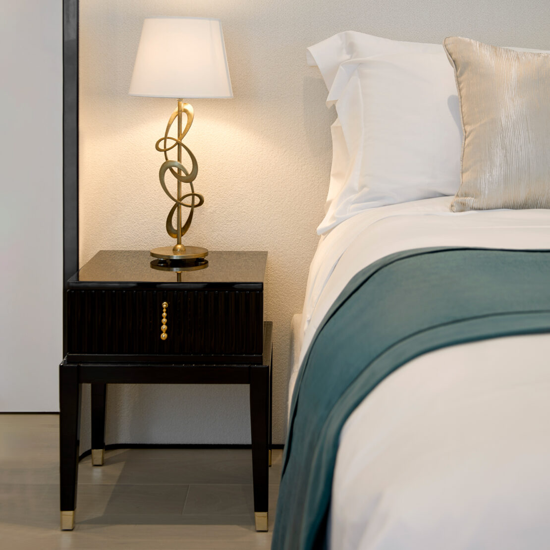 A modern bedroom showcases an Art Deco Style Bedside Table with golden accents beside a neatly made bed. The nightstand is adorned with a decorative table lamp featuring a twisted gold base and a white shade. The bed is dressed in pristine white linens, complemented by a teal blanket and a metallic accent pillow.