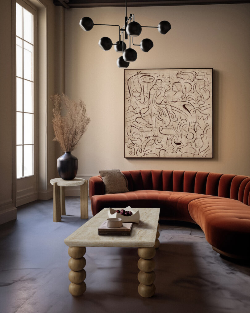 A modern living room with a curved red sofa, abstract wall art, a geometric chandelier, and a uniquely designed coffee table. A vase with dried branches sits on a side table