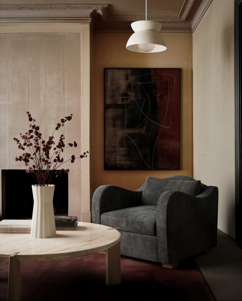 A modern living room with a dark gray armchair, an abstract painting on the wall, and a round table with a white vase of red foliage. A white pendant light hangs from the ceiling