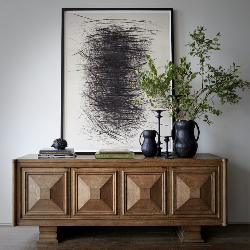 A wooden sideboard with geometric patterns holds a leafy plant, two black vases, books, and decorative items. A large abstract artwork is hung above it on the wall.