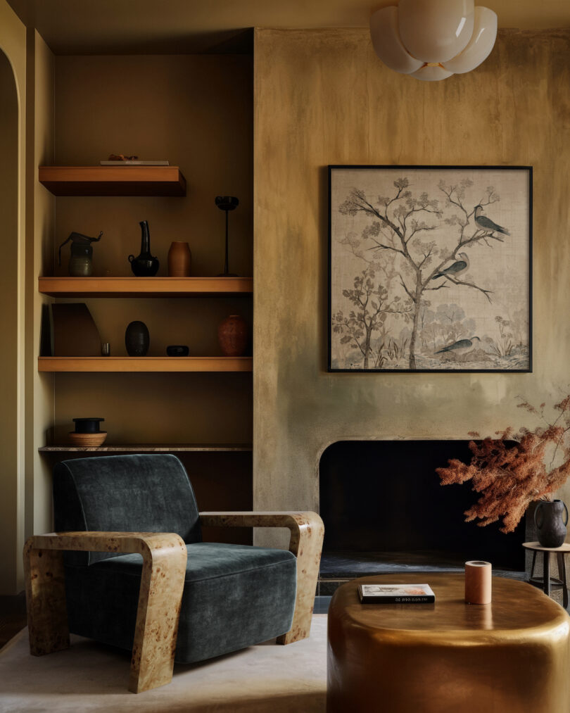 A cozy living room with a dark armchair, a modern coffee table, and shelves with decor. A framed bird illustration hangs above the fireplace. Warm, earthy tones adorn the space