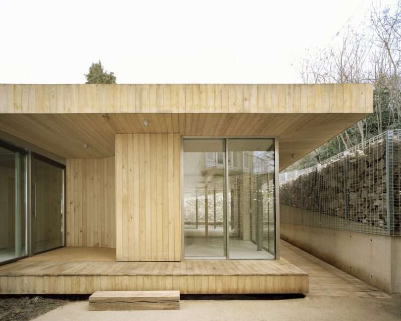 A modern wooden structure with large glass doors and windows, set on a light concrete base, surrounded by trees and a wire fence.