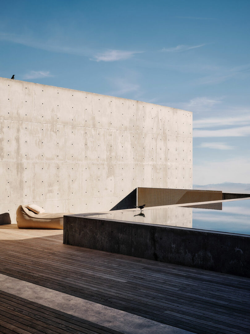 Modern architectural setting with a minimalist concrete wall, wooden deck, and an infinity pool reflecting the sky. A white lounge chair is placed on the deck.