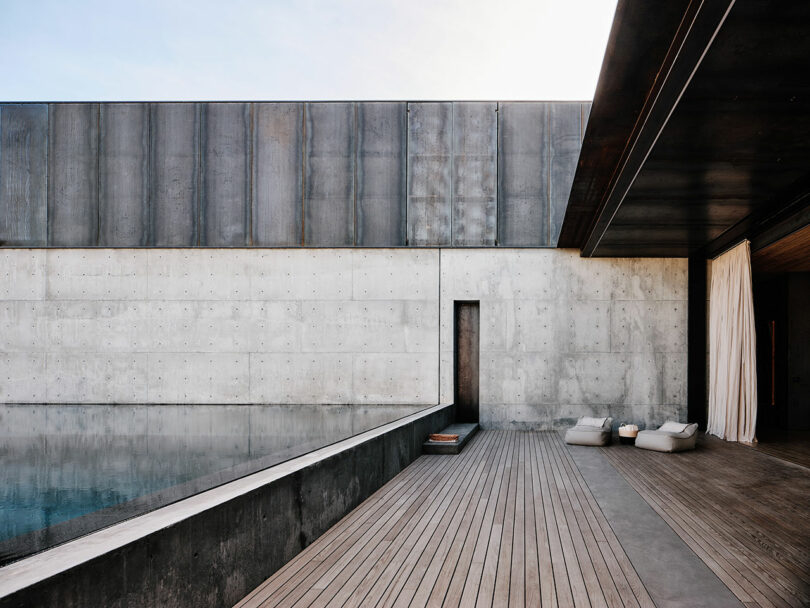 A modern exterior features a wooden deck, a reflecting pool, and concrete walls with a single narrow door. Cushions and a yoga mat are placed on the deck beside the pool.