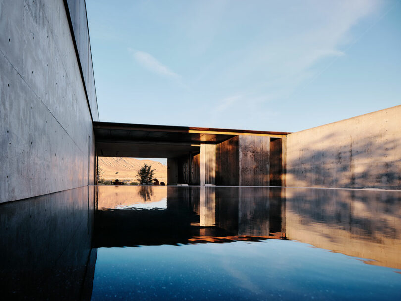 Rectangular outdoor pool next to modern concrete architecture with open-air sections, reflecting the blue sky and surrounding landscape in the water's surface.