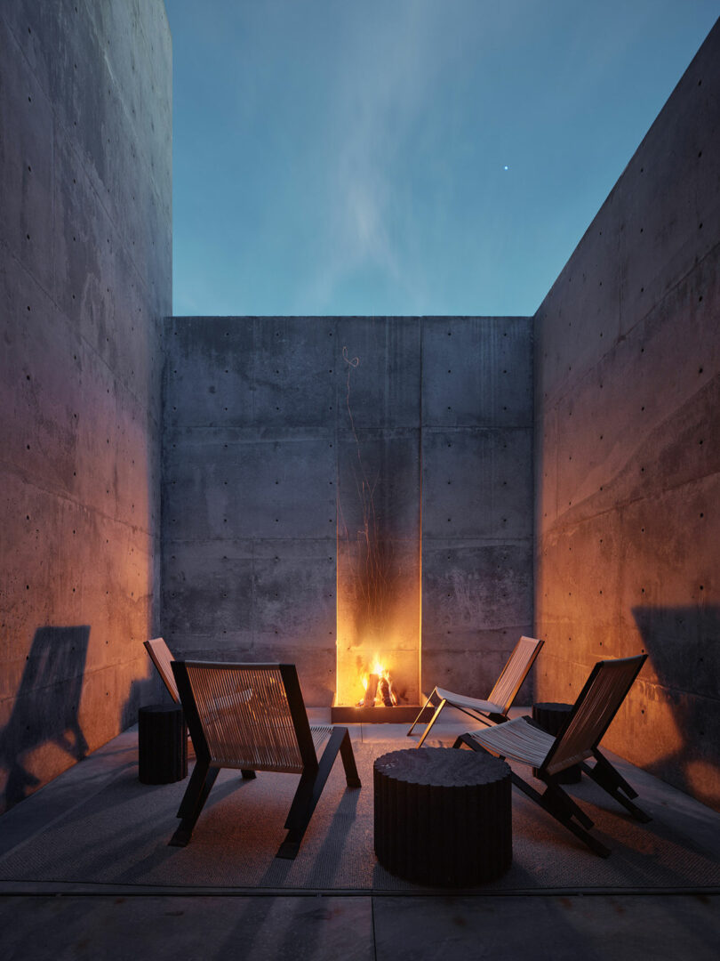 A modern outdoor space with concrete walls, featuring four lounge chairs around a rectangular fire pit, illuminated by the fire's glow. The sky is twilight blue.