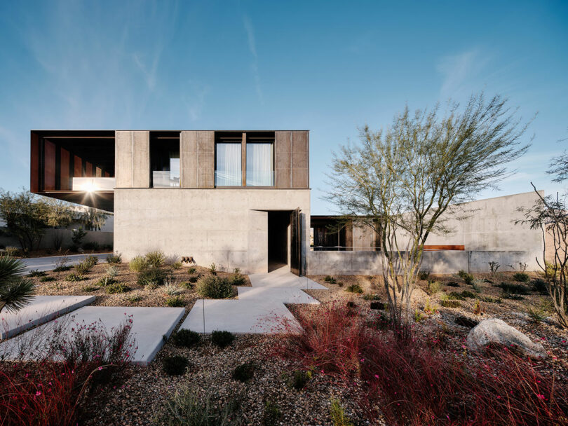 Modern two-story house with large windows and wooden accents set in a desert landscape with minimalistic pathways and xeriscaping.