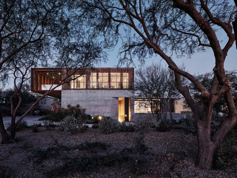 Modern two-story house with large windows and a blend of concrete and wooden elements, surrounded by trees and natural landscaping, photographed at dusk.