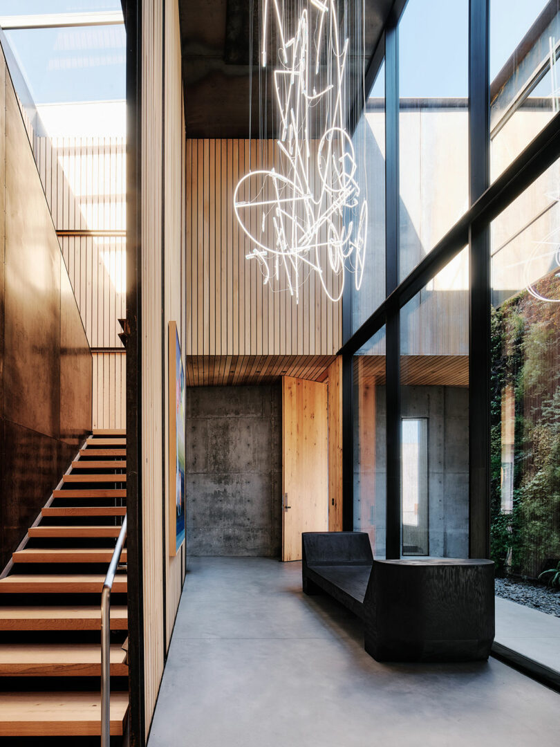 Modern interior with concrete walls, wooden paneling, large windows, a black bench, and illuminated abstract light fixture. A staircase with a metal railing leads to an upper level.