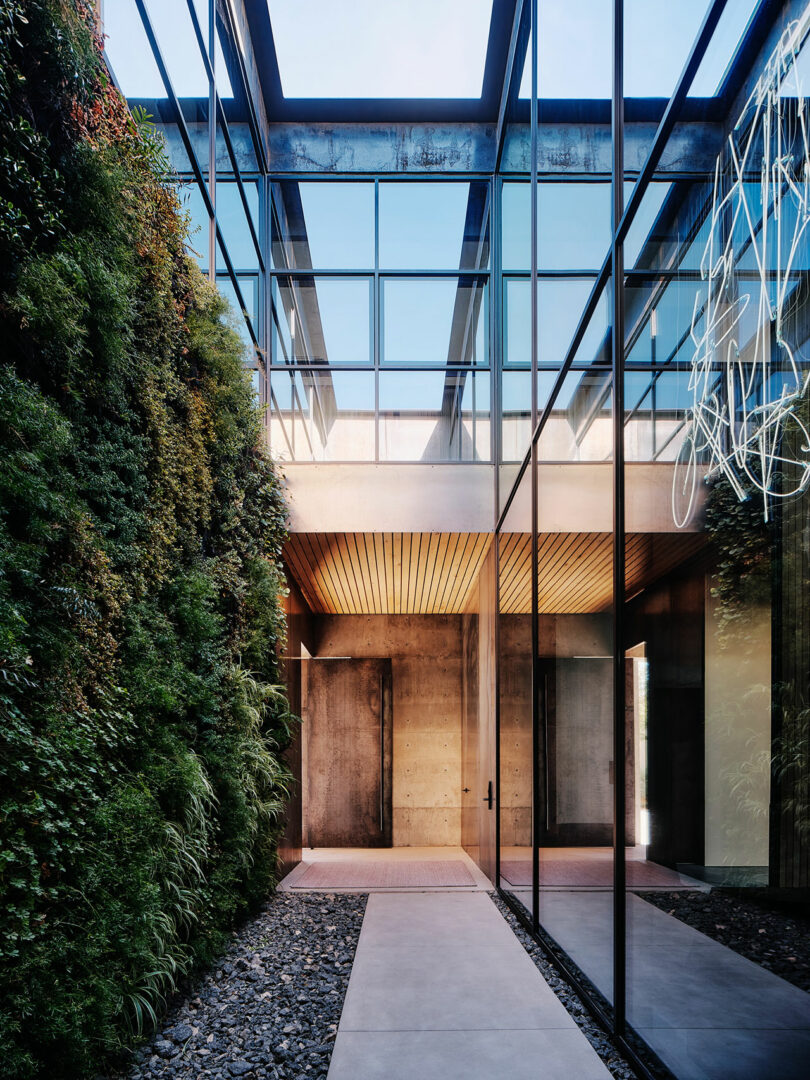 Modern architectural entryway featuring a glass ceiling, a vertical garden wall on the left, and large glass windows on the right. The space leads to a wooden door at the far end.