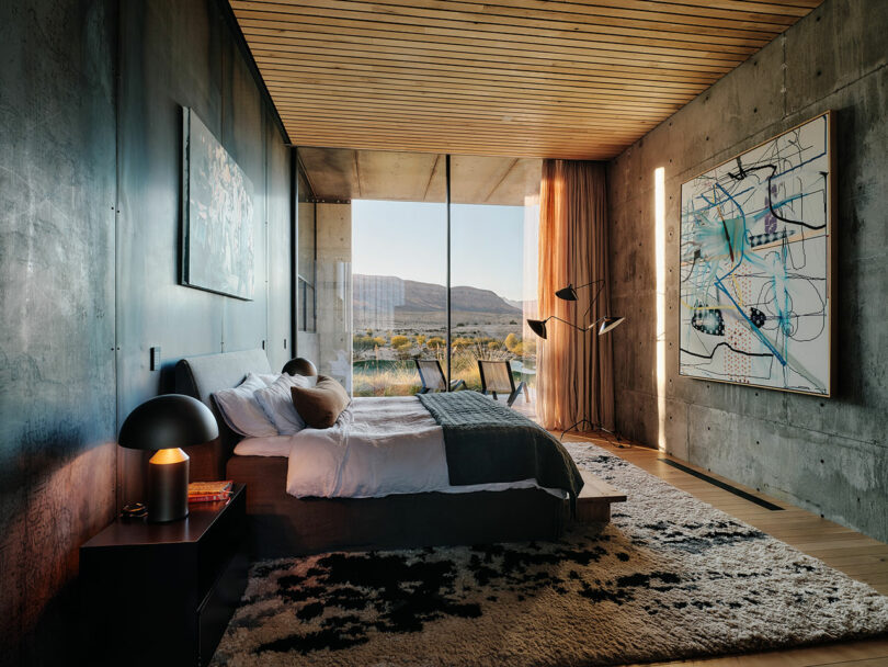 Modern bedroom with large floor-to-ceiling windows, a double bed, contemporary art on the walls, and minimalist furnishings. View of a mountainous landscape outside.