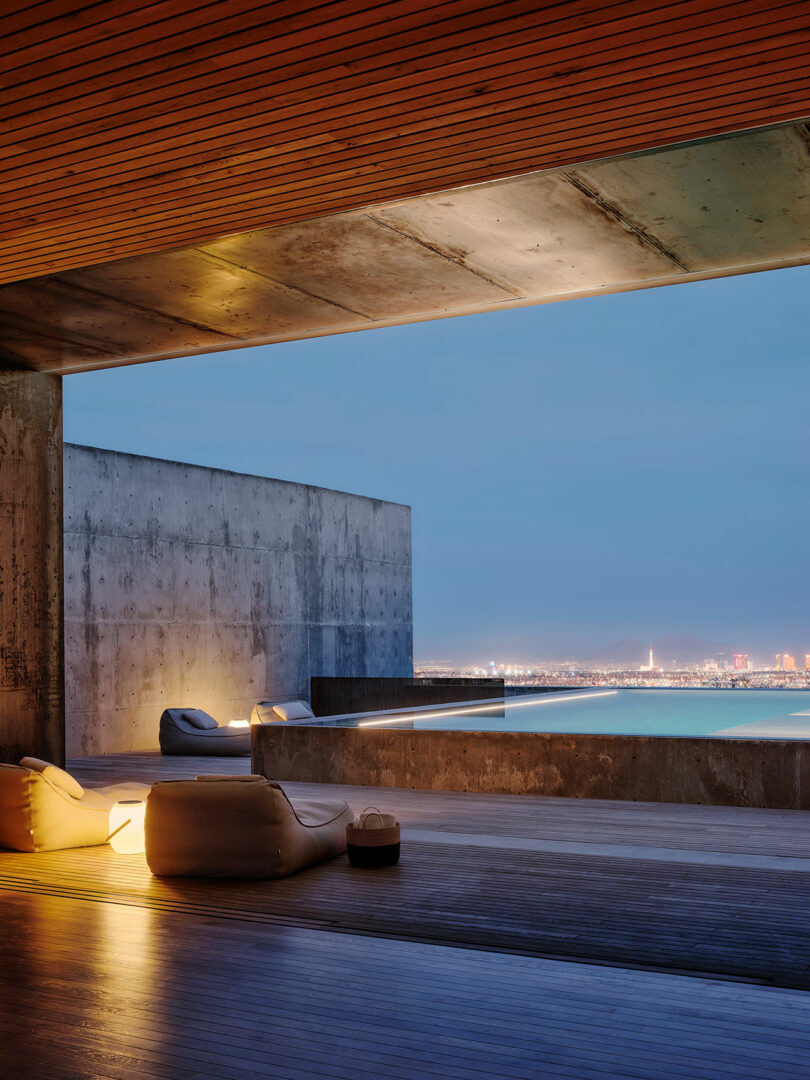 Modern concrete balcony with lounge seating, ambient lighting, and a view of a distant city skyline and waterfront at dusk.