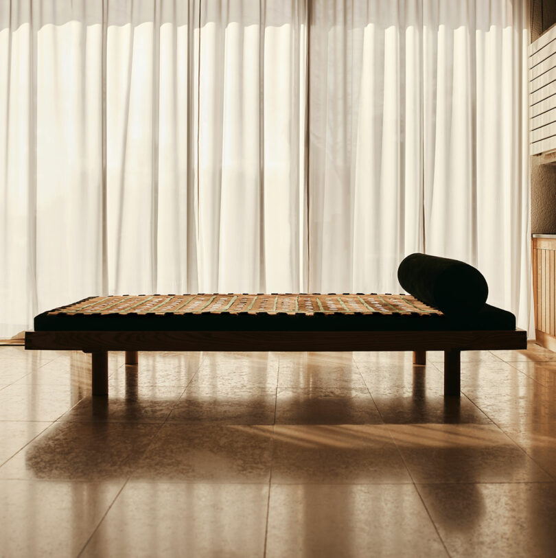 A minimalist wooden daybed by Yves Salomon with a dark mattress and cylindrical pillow is placed in front of tall, sheer white curtains in a sunlit room.