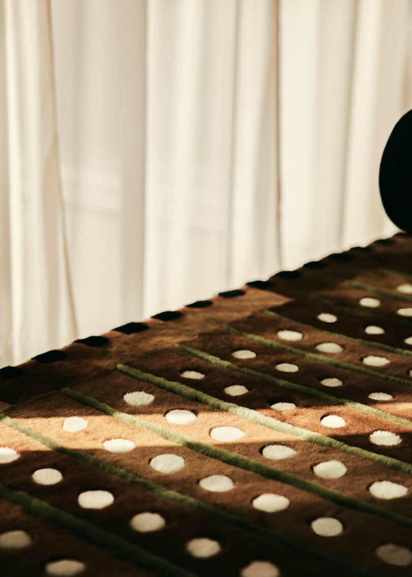 A patterned carpet by Yves Salomon with a brown and green striped design featuring white dots, against a backdrop of white curtains.