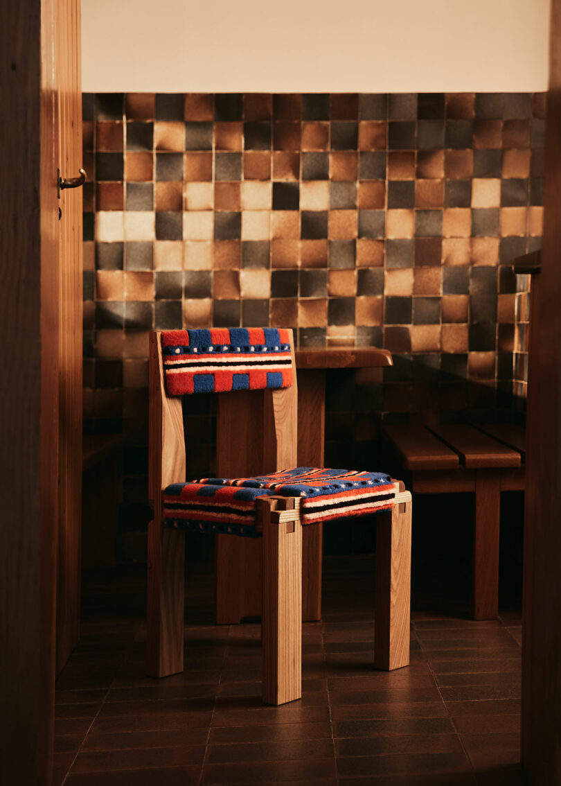 A wooden chair by Yves Salomon with colorful striped fabric cushions is placed in a room with brown tiled walls and a brown tiled floor.