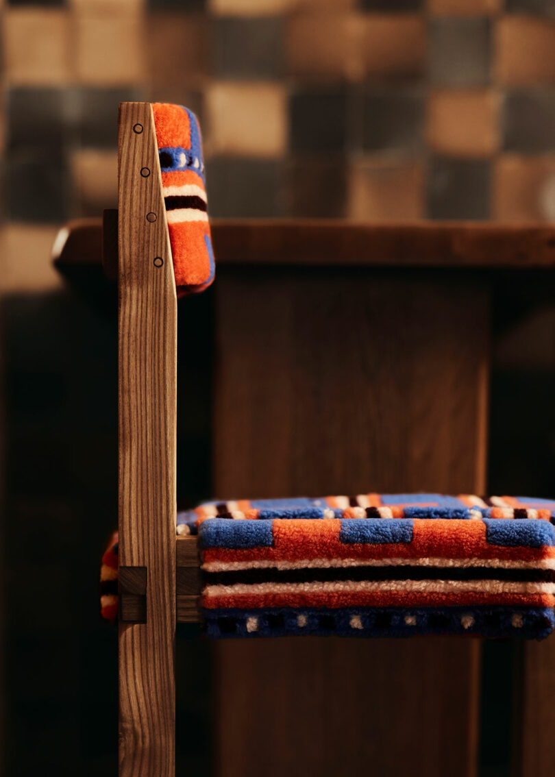 A wooden chair by Yves Salomon with a colorful woven cushion, positioned next to a wooden table with a blurred checkered background.