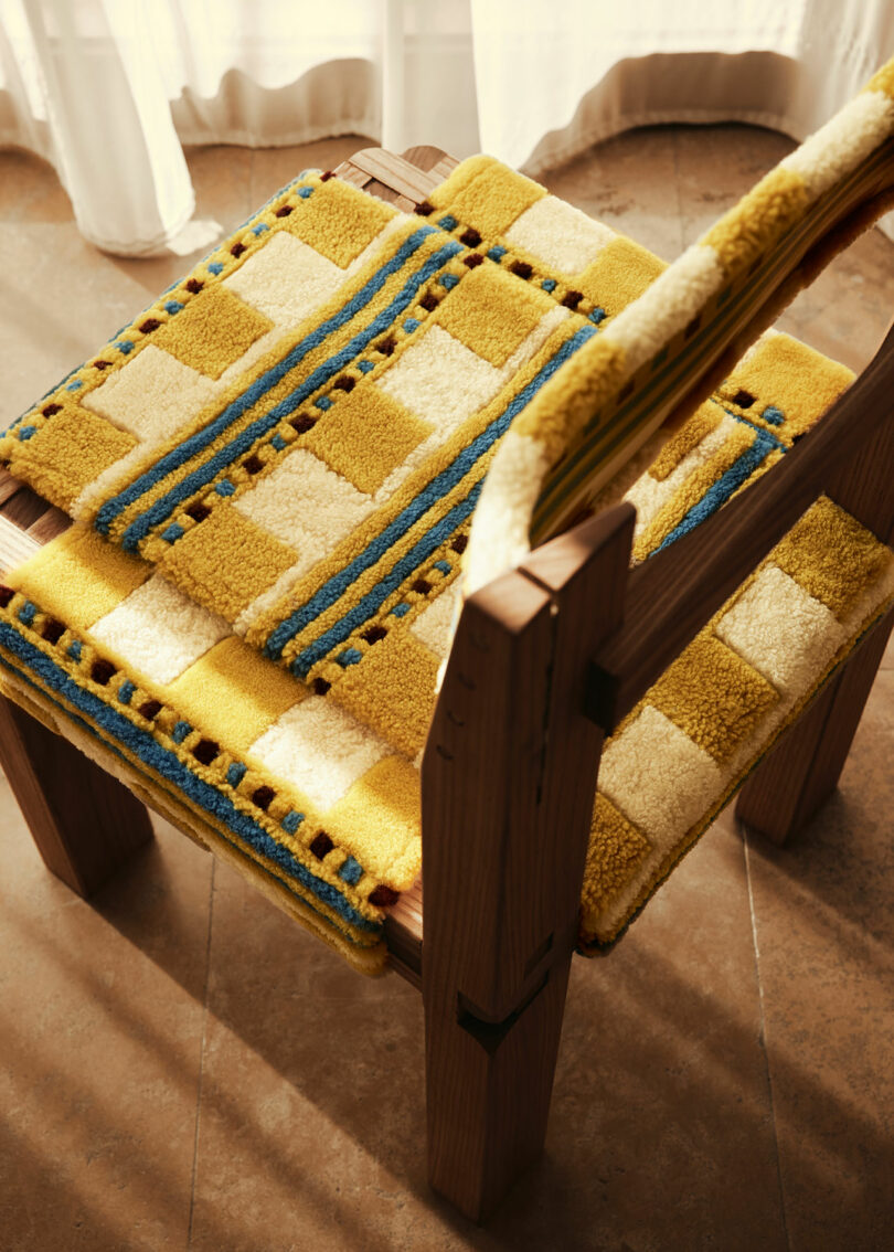 A wooden chair by Yves Salomon features a yellow, white, and blue checkered cushion and backrest cover. Sunlight casts shadows on the tiled floor. White curtains are partially visible in the background.