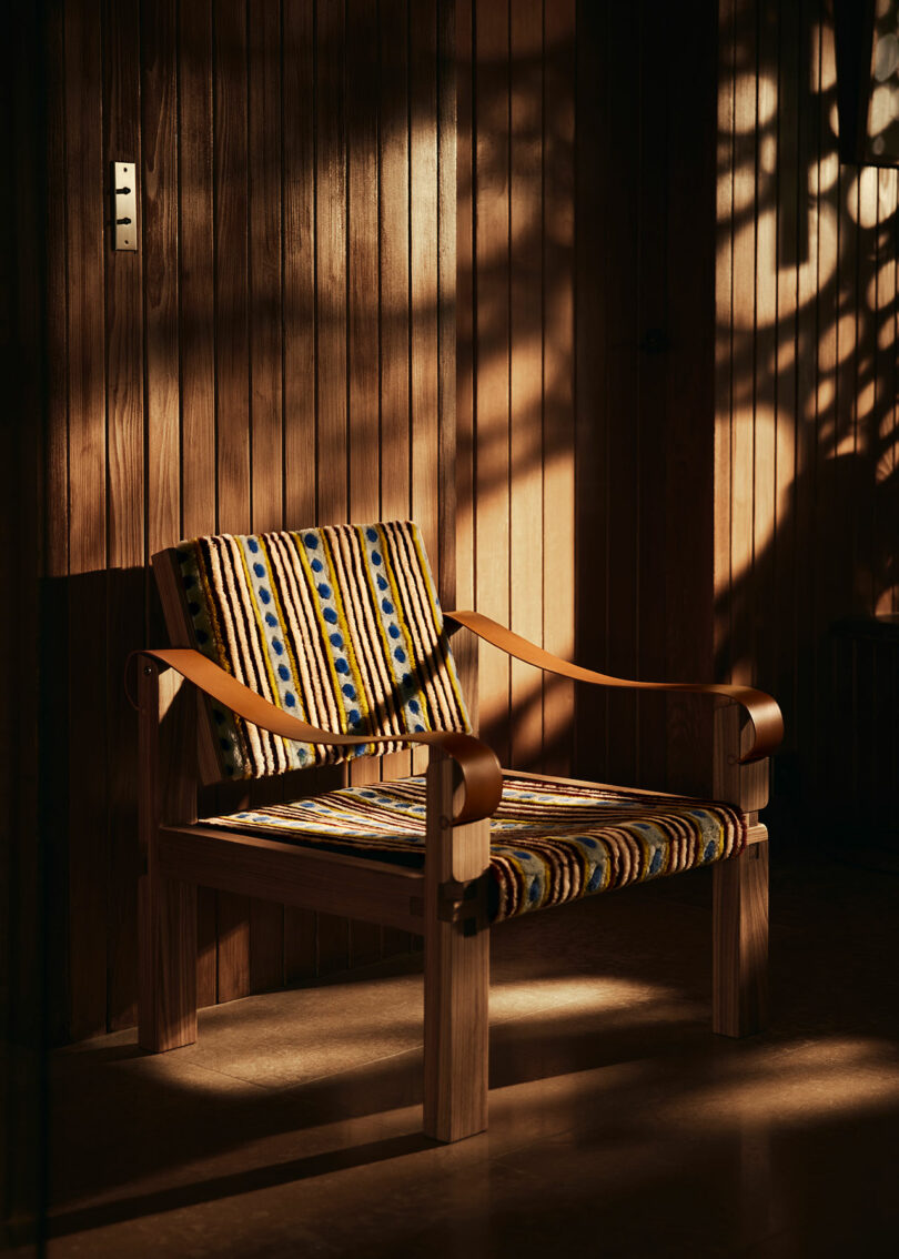 A wooden chair by Yves Salomon with patterned upholstery is placed against a wooden paneled wall. Sunlight casts shadows on the wall and the chair.