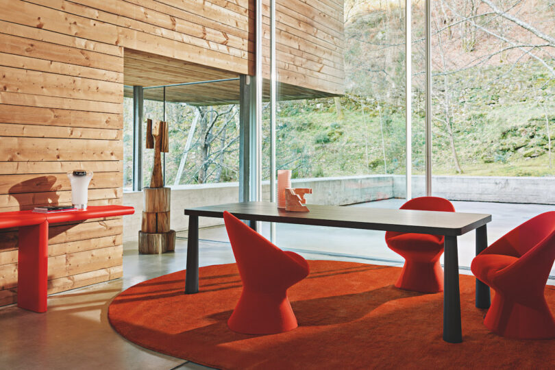 A modern indoor space featuring a wooden interior, red furniture, a black table, and floor-to-ceiling windows overlooking a serene outdoor area