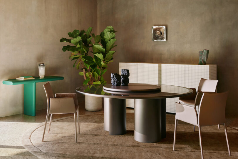 A modern room with a round table and chairs, a green side table with a vase, a large plant, and a white cabinet on a textured rug. A small framed picture is mounted on the beige wall