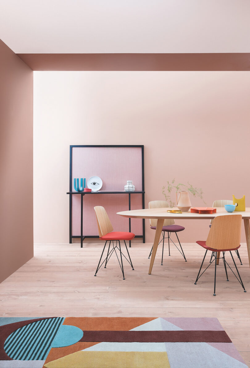 A modern dining room with a round wooden table, four colorful chairs, and a decorative console in the background. The room features light wood flooring and soft pink walls