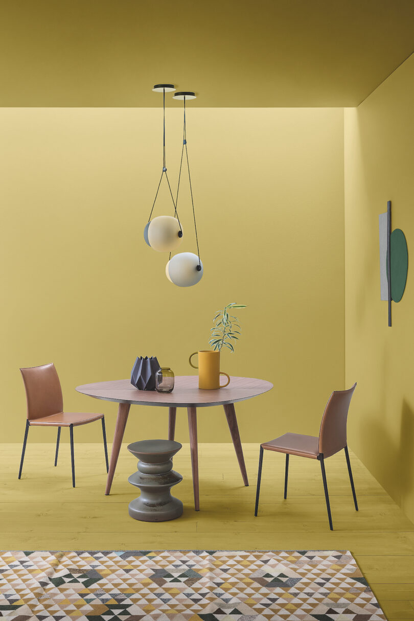 A modern dining setup with a wooden table, three brown chairs, a geometric-patterned rug, and spherical pendant lights against a yellow background. A yellow pitcher and a small plant are on the table