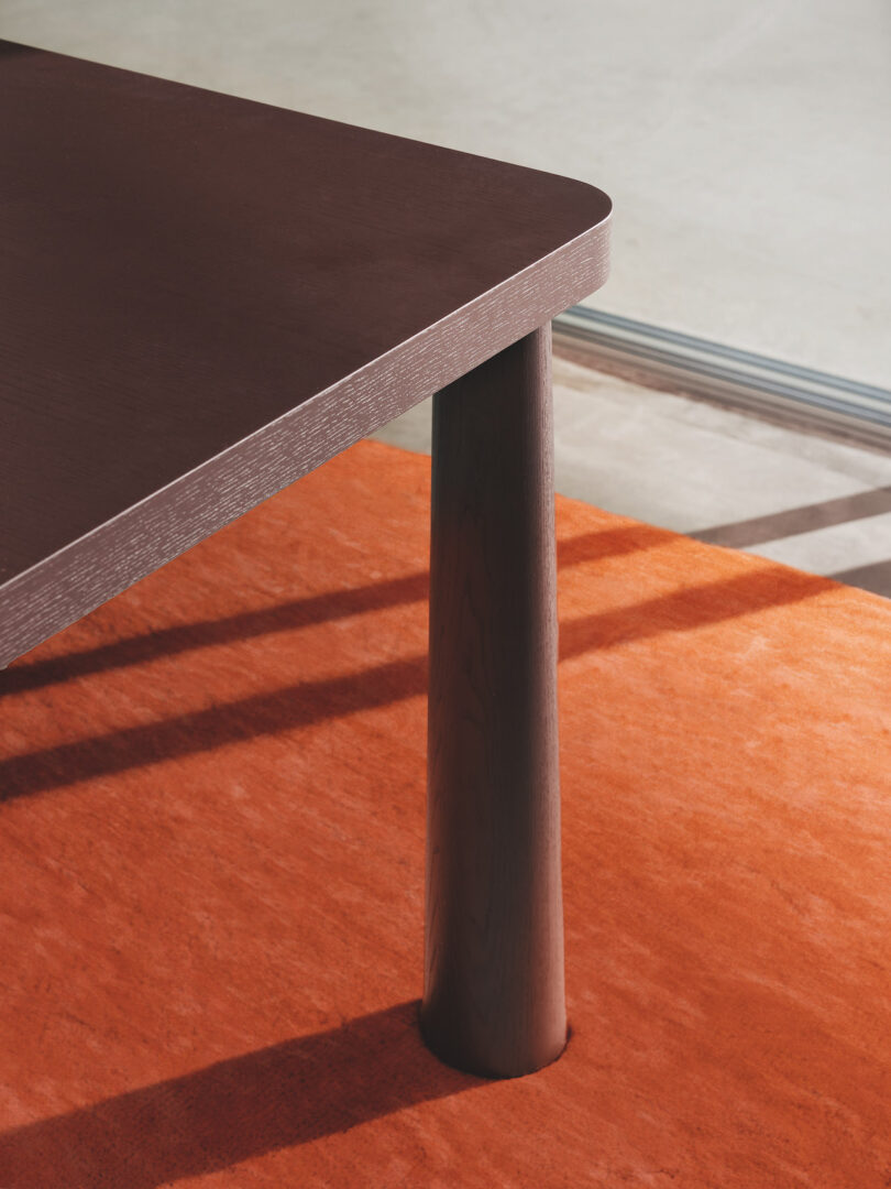 Corner of a brown wooden table with rounded edges, standing on an orange carpet with sunlight casting shadows