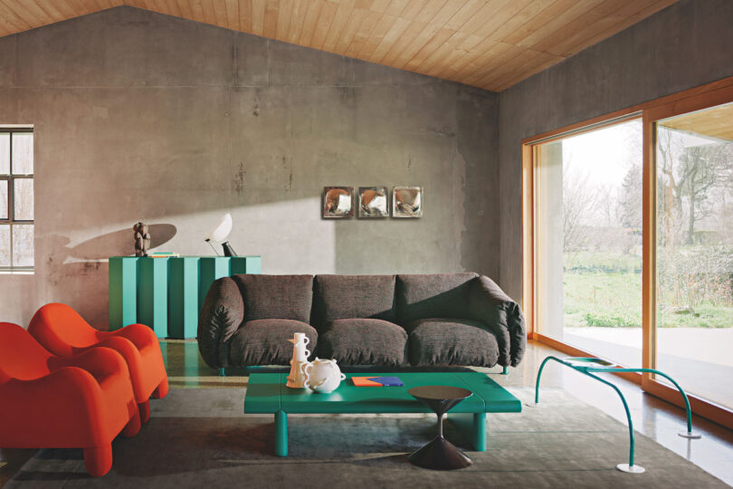 Modern living room with concrete walls, large windows, and wooden ceiling. Features a gray sofa, two orange chairs, green coffee table, and a green console table