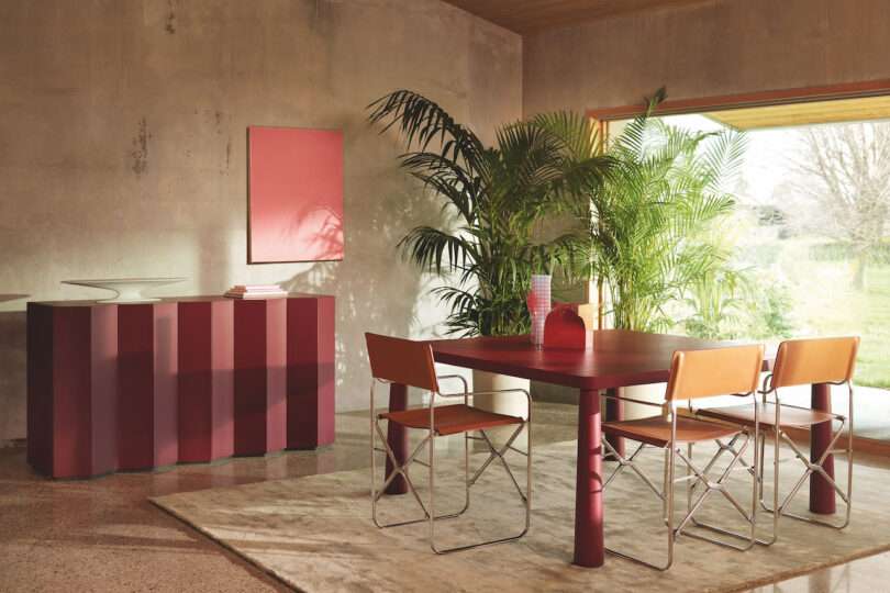 A modern dining area with a red table and four brown chairs, a sideboard, indoor plants, and a large window on the right providing natural light. The walls have a concrete finish