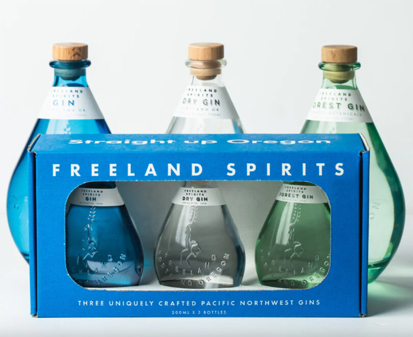 A set of six uniquely shaped bottles, three in a blue box labeled "Freeland Spirits," and three in the background. Each bottle has a cork stopper and clear labeling.