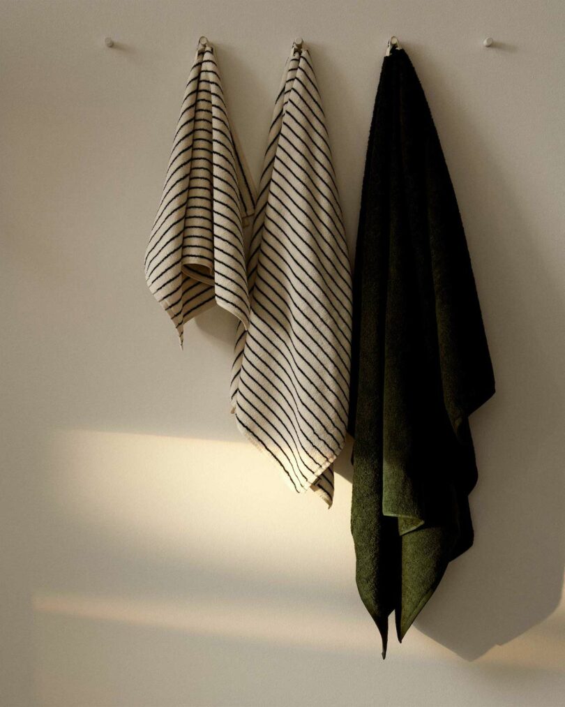 three towels hanging on a wall; from left to right: a white with green stripes hand towel, a white with green stripes bath towel, and an all dark green bath sheet.