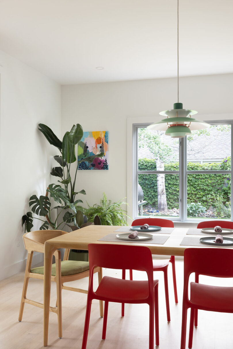 Modern dining room with a wooden table, red chairs, a pendant light, and plants by a large window. A colorful painting is on the wall.