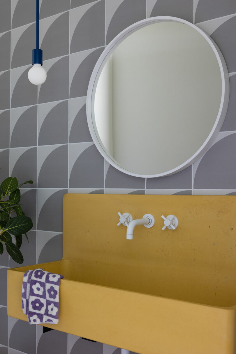 A bathroom features a round mirror, geometric gray tiles, a yellow sink with two knobs, a white faucet, a hanging bulb, and a purple and white patterned towel. A plant is on the left.