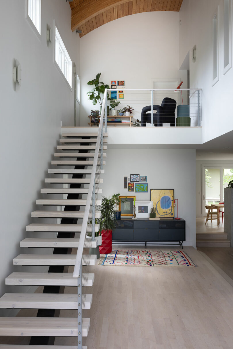 Modern interior with a white staircase, art on the walls, and plants. A black cabinet with decor underneath a loft area with chairs and a plant. Natural light fills the space.