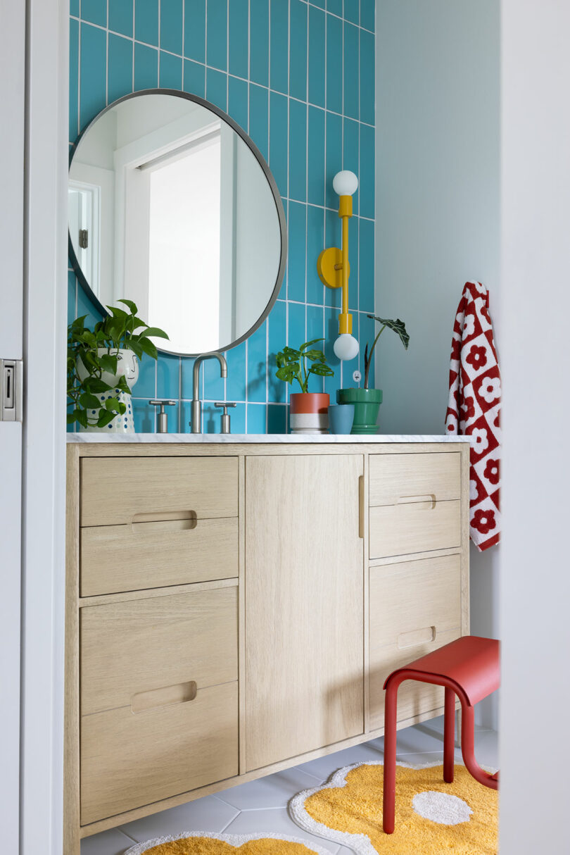 Bathroom with blue tiled walls, round mirror, light wood vanity, plants, and colorful decor, including a red stool and a yellow patterned rug.