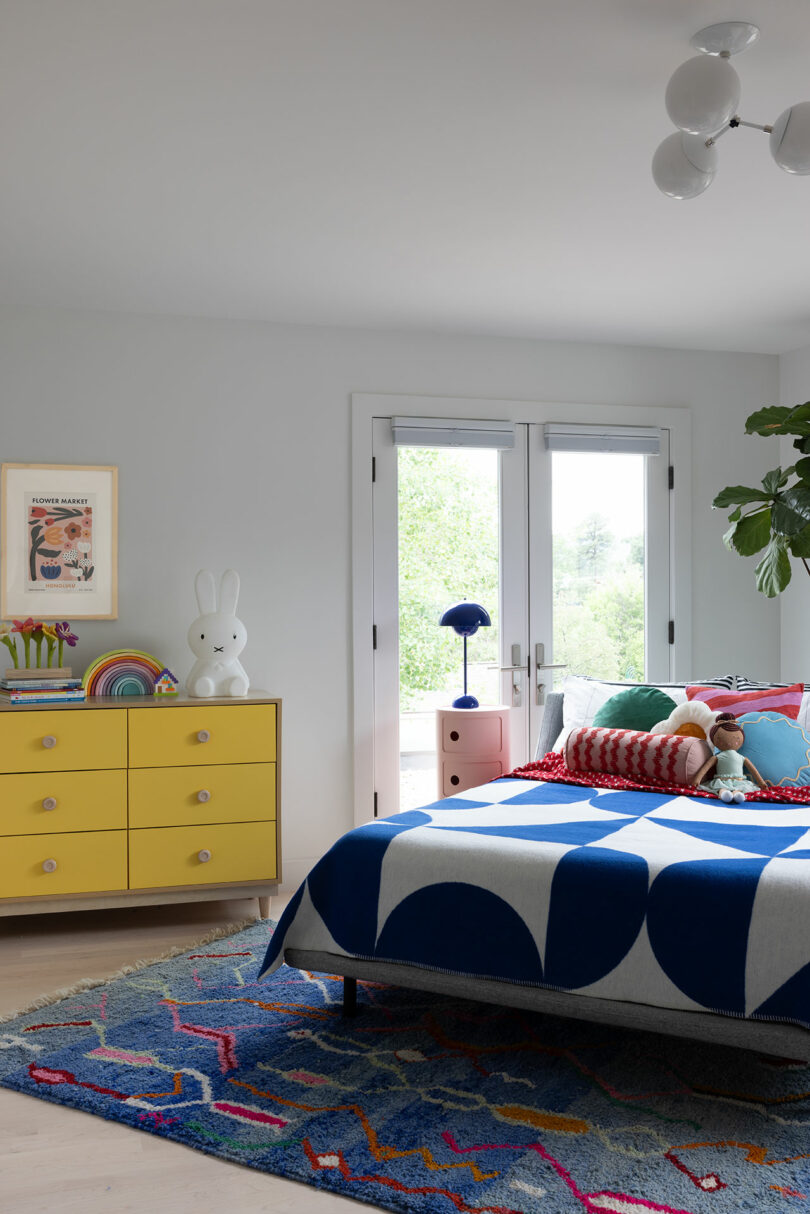 Bright bedroom with a blue and white bedspread, colorful cushions, a yellow dresser, and playful decorations, including a white rabbit lamp. Large windows let in natural light.