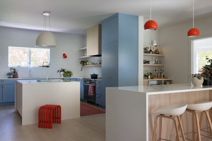 Modern kitchen with blue cabinets, white countertops, and pendant lights. Includes a central island, red stool, and shelves with various items. Bright, natural light streams through the window.