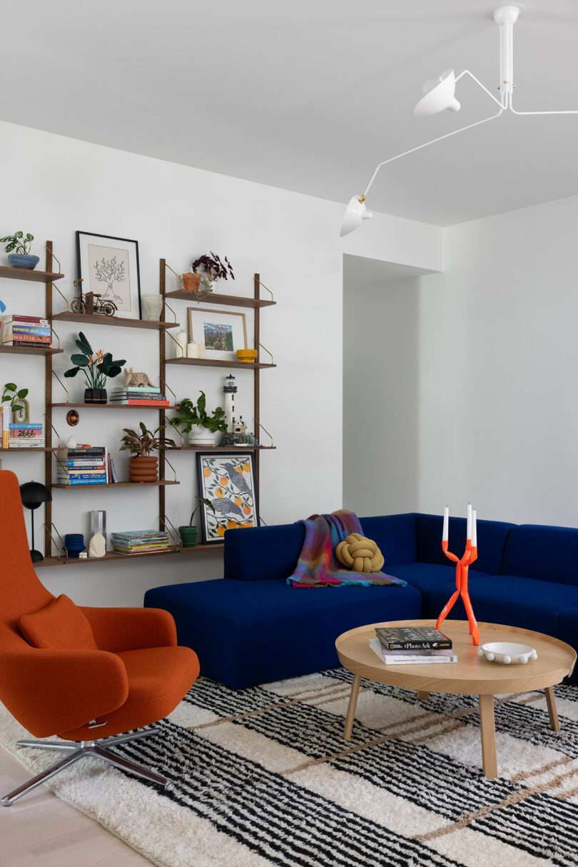 A modern living room with a blue sectional sofa, orange chair, round wooden coffee table, and a wall-mounted bookshelf. A black and white patterned rug covers the floor.