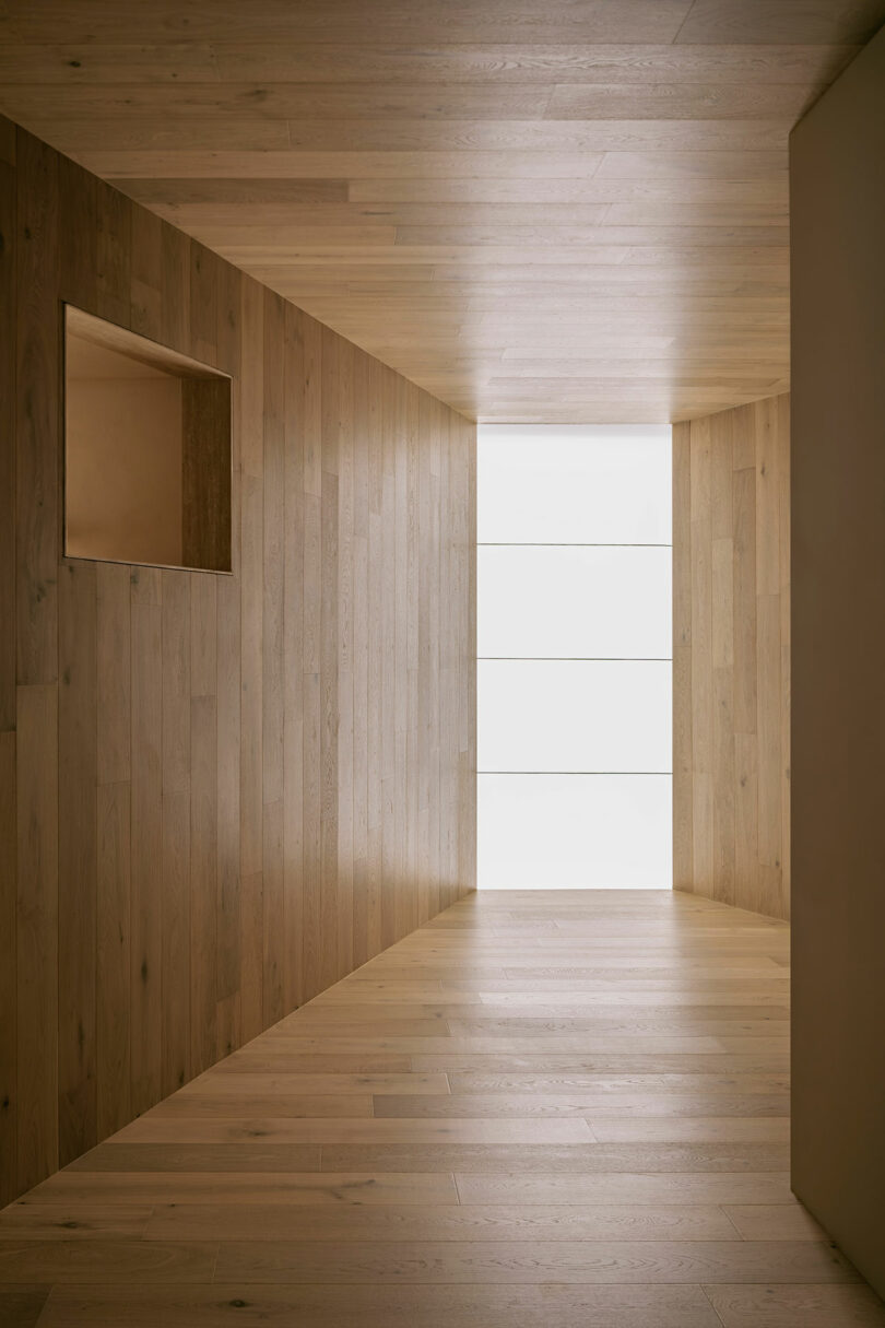 A narrow, light-filled hallway with wooden walls and flooring. A square window is set into the left wall, and the far end is illuminated by soft, natural light.