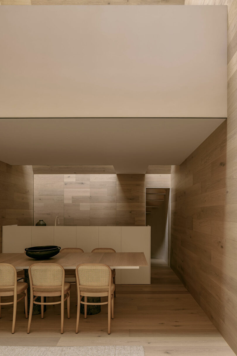 Minimalist dining area with a wooden table, six matching chairs, neutral-toned walls, and a modern kitchen in the background.