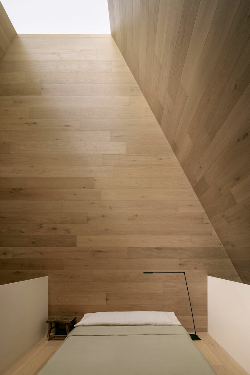 Minimalist bedroom with angled wooden walls, a low bed with a green cover, and a slender black lamp.