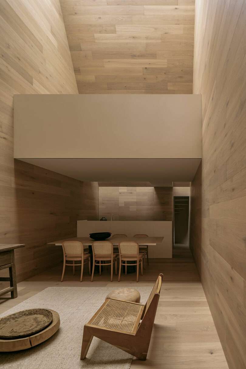 Minimalist interior with wooden walls, featuring a dining area, table, six chairs, and a low wooden chair in the foreground. Natural light streams from a high window.