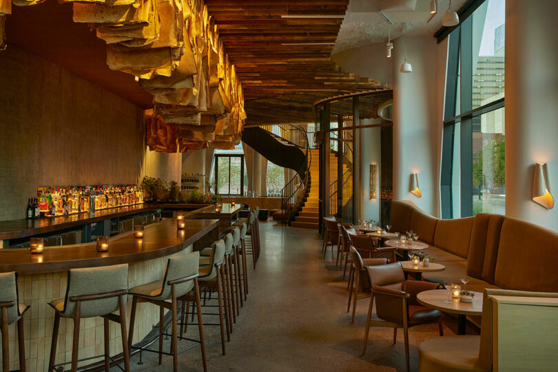 A modern, dimly lit bar with wooden decor, featuring a long counter with stools, tables with chairs, and a spiral staircase in the background.